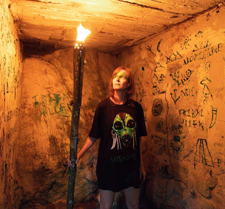 Woman inside a cave holding a torch, wearing a Losaquis tribal alien t-shirt, surrounded by ancient pictograms, symbolizing alien identity and ancestral art.
